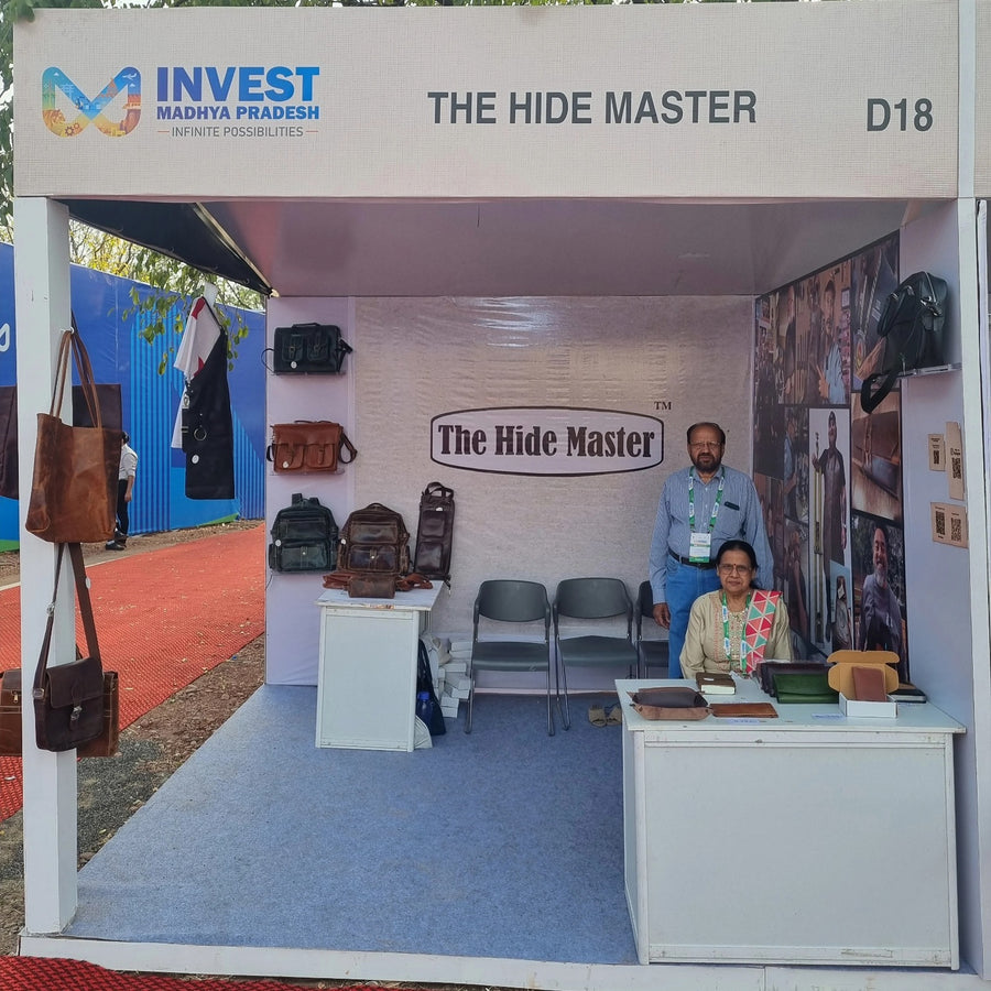 A booth display for 'The Hide Master' at a fair or exhibition, featuring a white structure with a blue floor, a branded sign, chairs, a desk, and two people standing and sitting inside. Bags and clothing are displayed on the left and right sides of the booth.