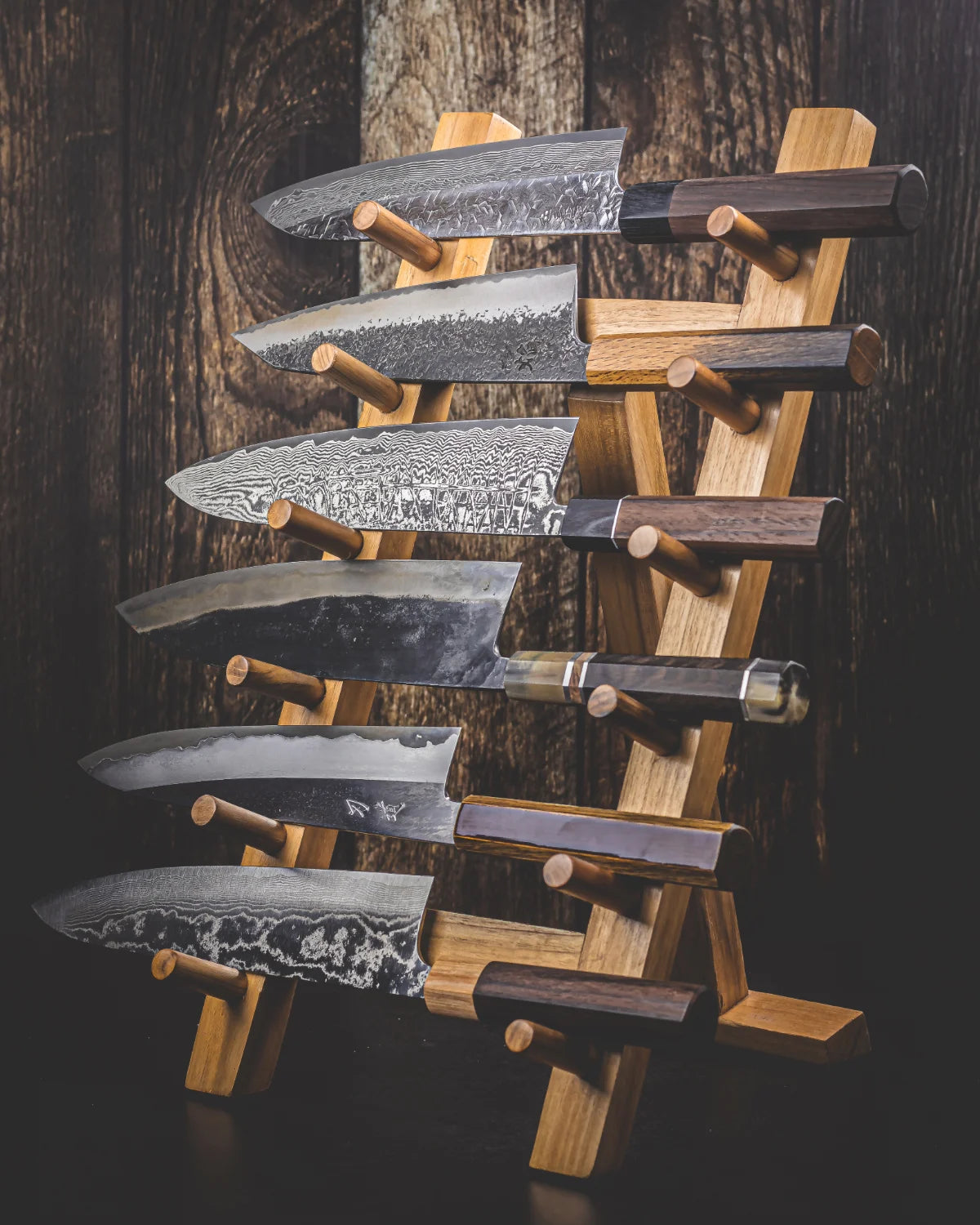 A set of six Japanese kitchen knives with Damascus steel blades and dark wood handles, displayed on a wooden knife tower rack stand against a dark wood plank background.